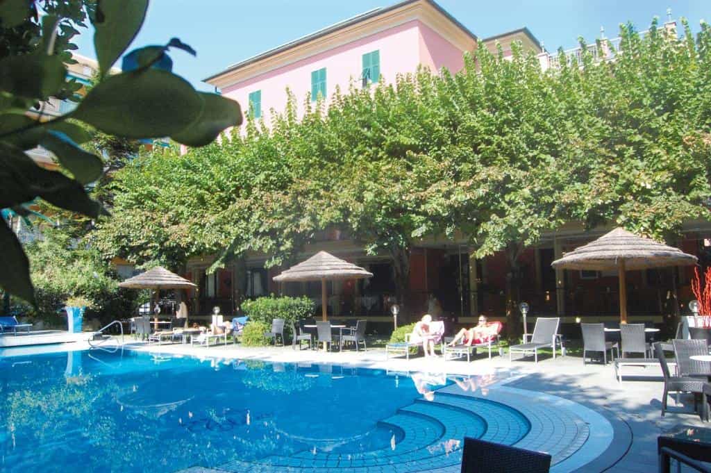 Swimming pool area with sun loungers and thatched umbrellas next to a pink hotel building.