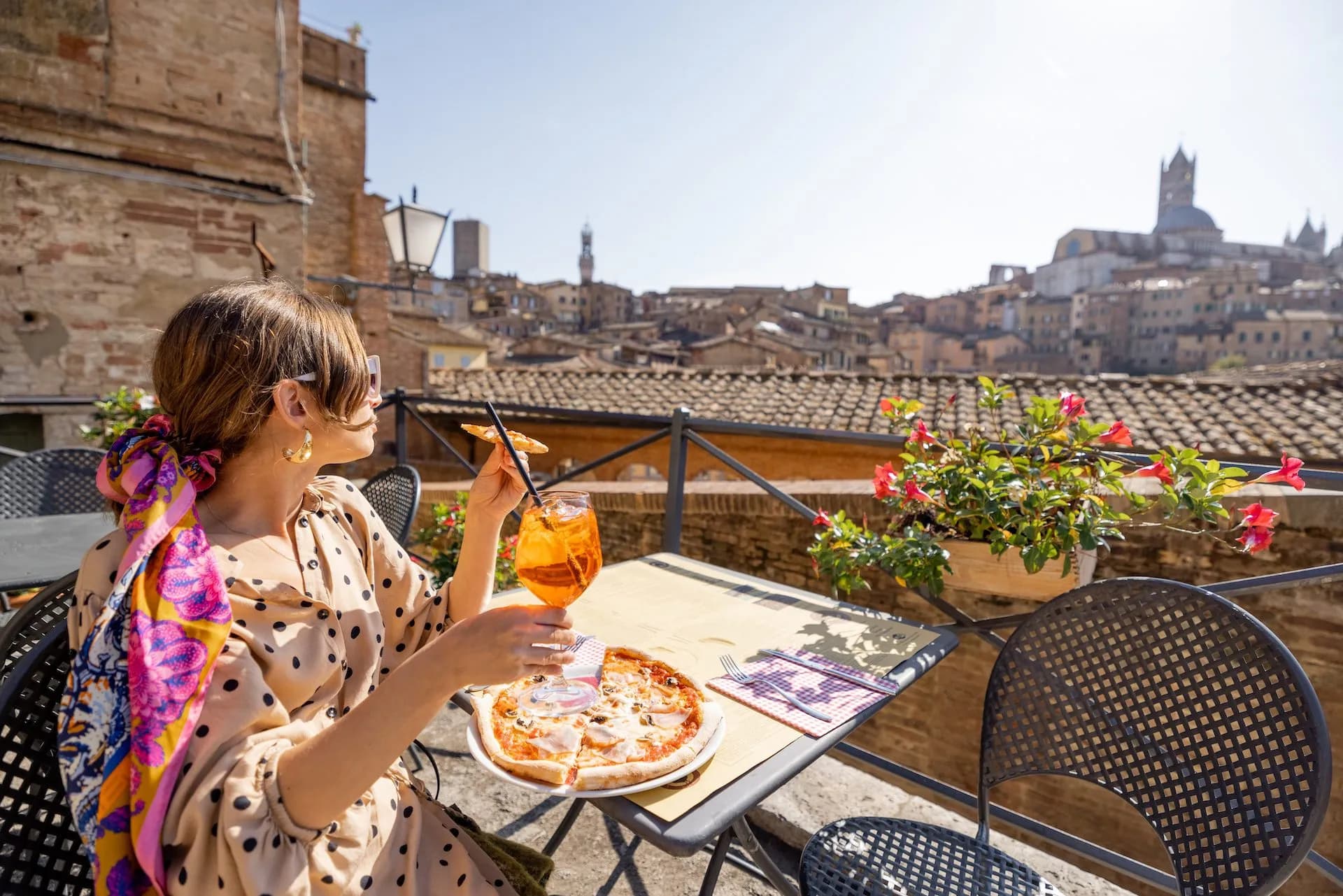 Woman dining on pizza and orange cocktail overlooking Siena cityscape and Duomo.