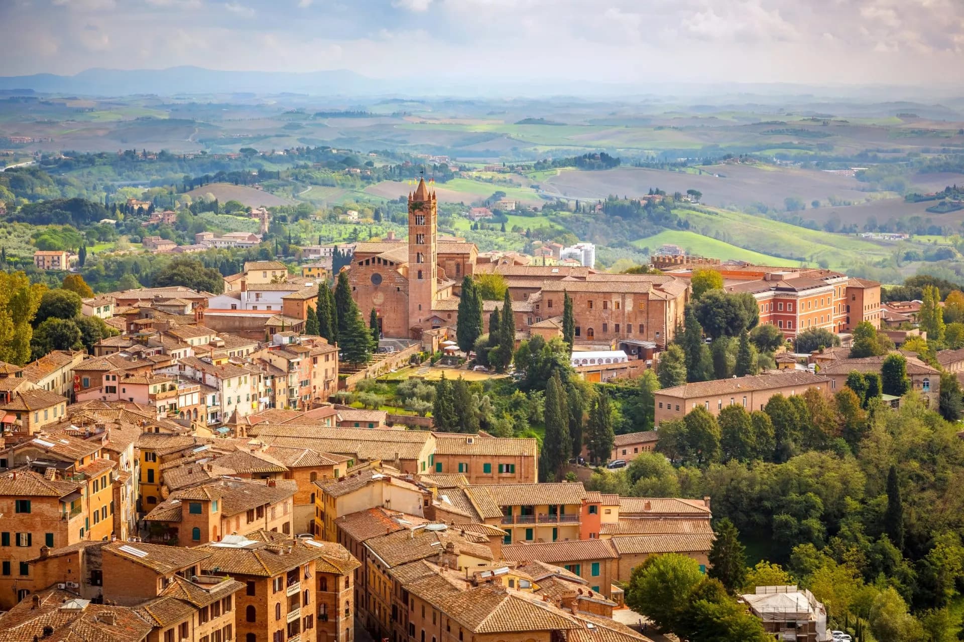 Aerial view of Siena's terracotta rooftops and historic buildings overlooking rolling Tuscan hills.