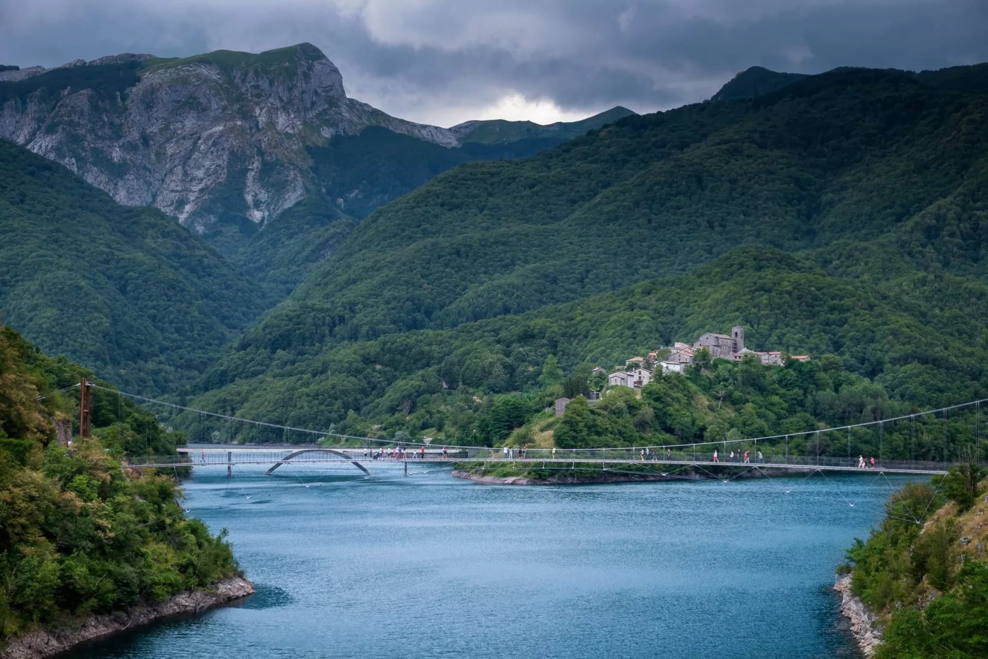 Suspension bridge over blue water at Lago di Vagli with village and forested mountains.