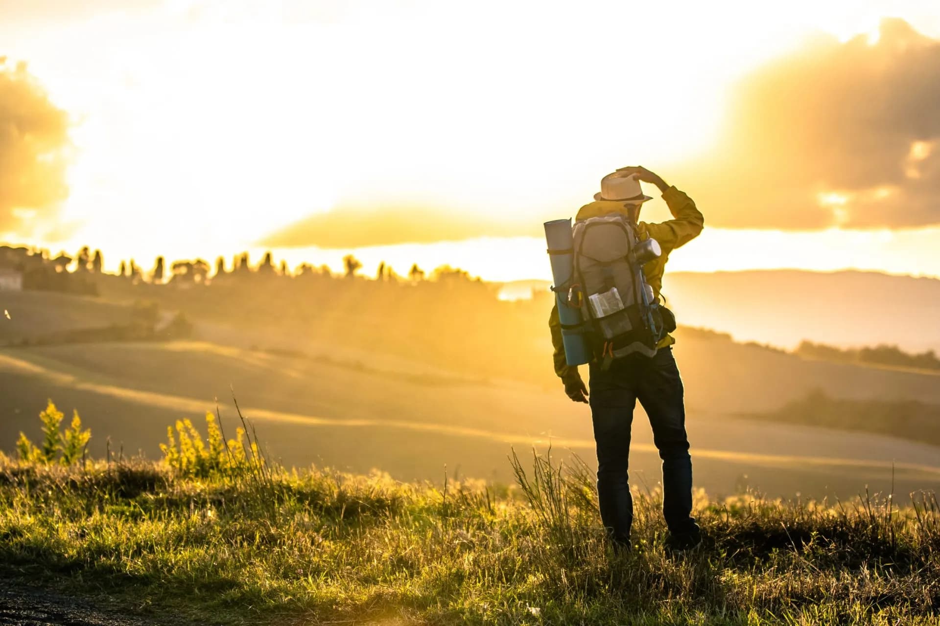 Hiker with backpack standing on grassy hill during sunset in Tuscany landscape