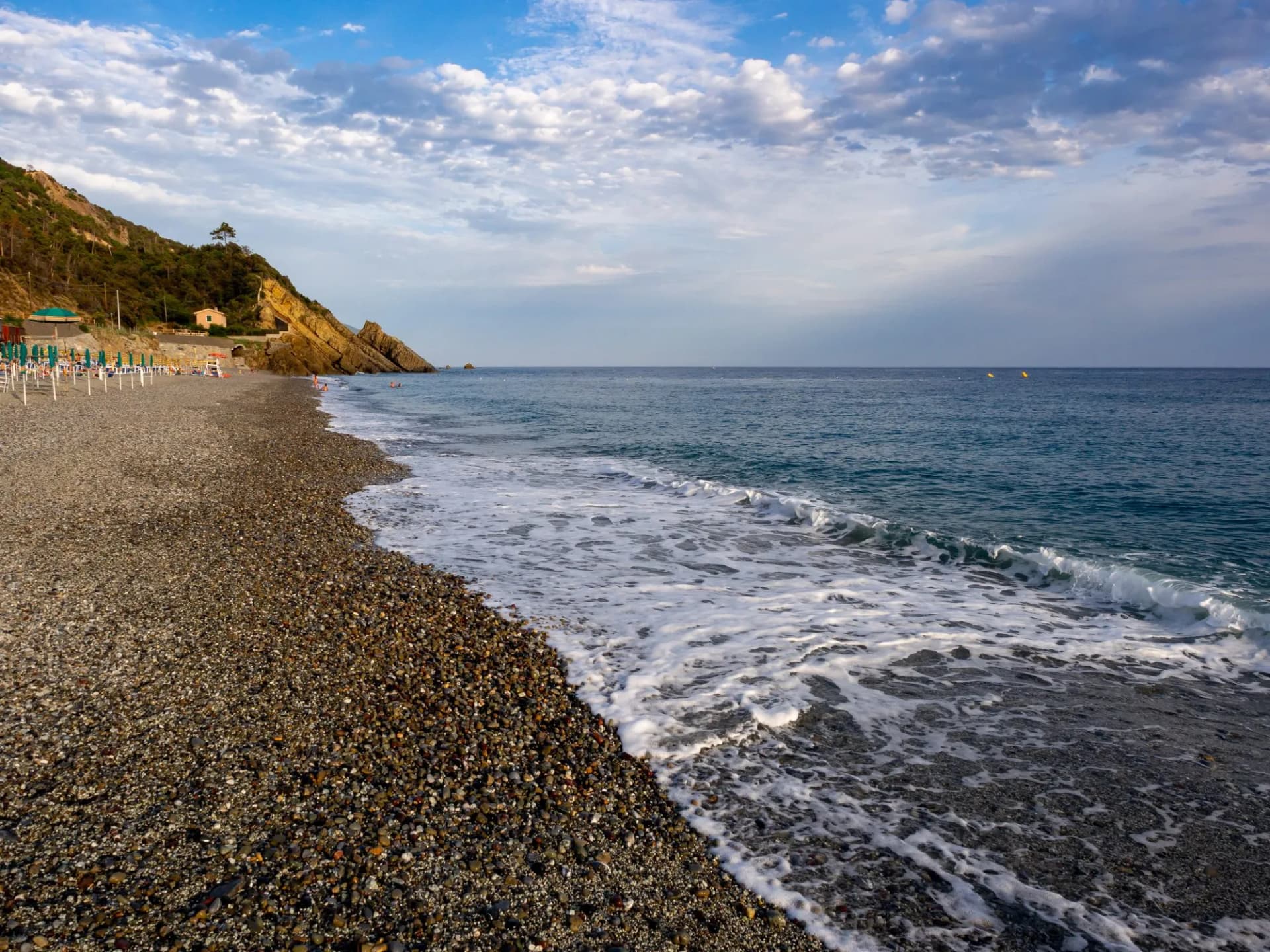 Pebble beach with waves meeting the shore, backed by a steep, green hillside in Deiva Marina.