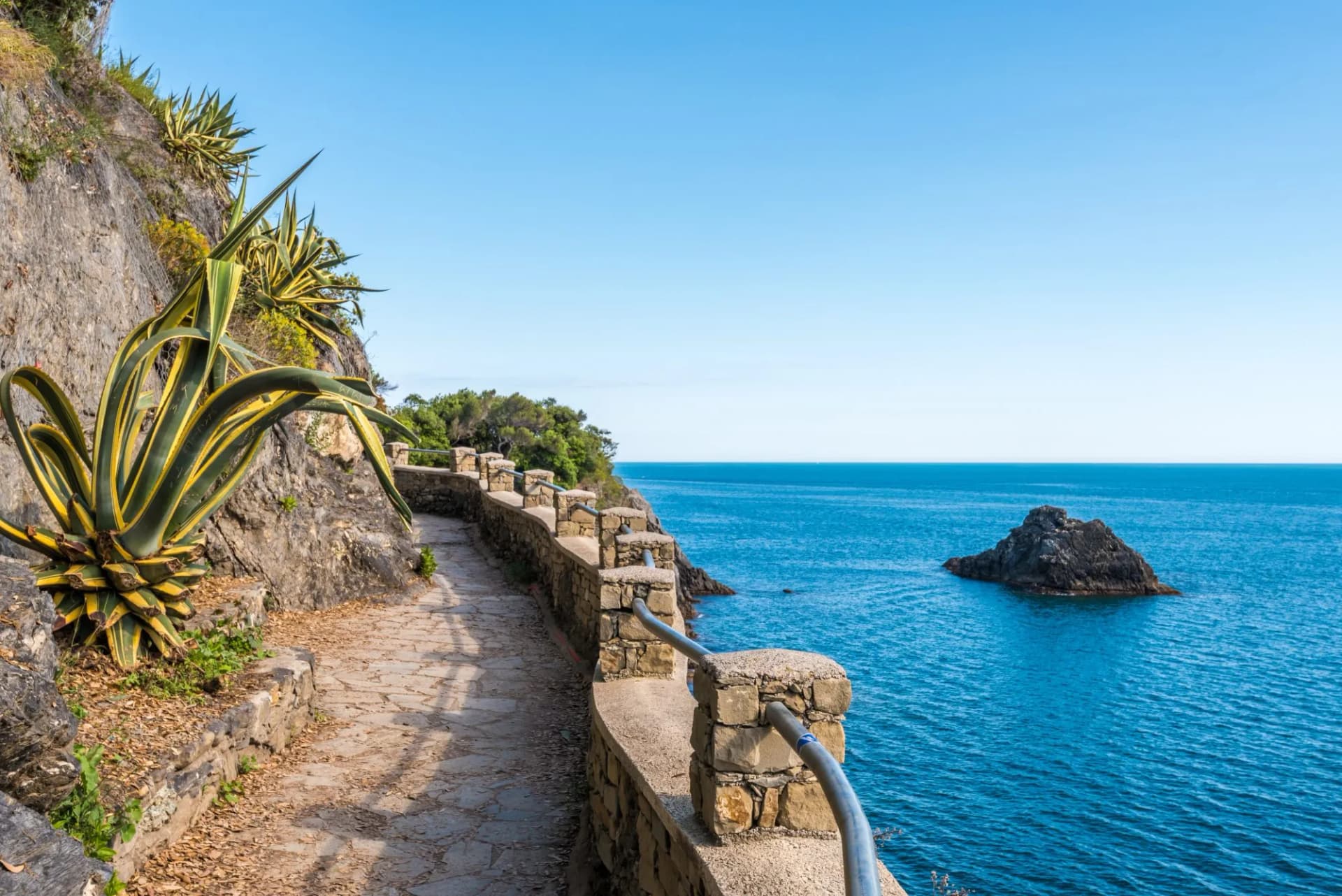 Coastal footpath with stone railing overlooking bright blue sea and rock formation, Ligurian Coast