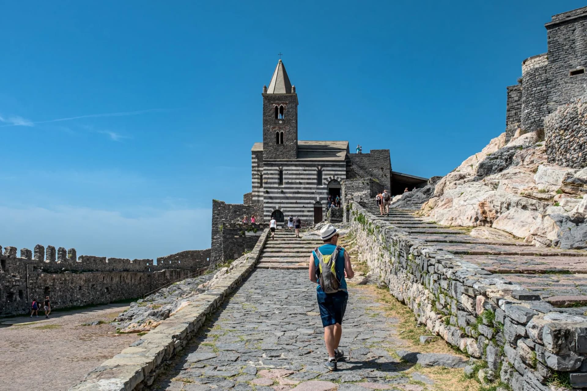 Hiker ascending stone steps toward Chiesa di San Pietro in Portovenere under a clear blue sky.