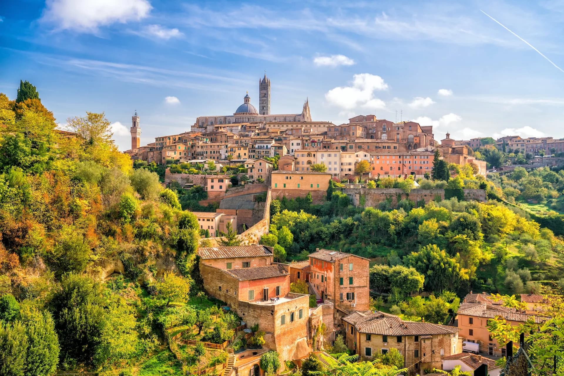 Historic hilltop city of Siena with terracotta buildings nestled in lush green and yellow foliage under a blue sky.