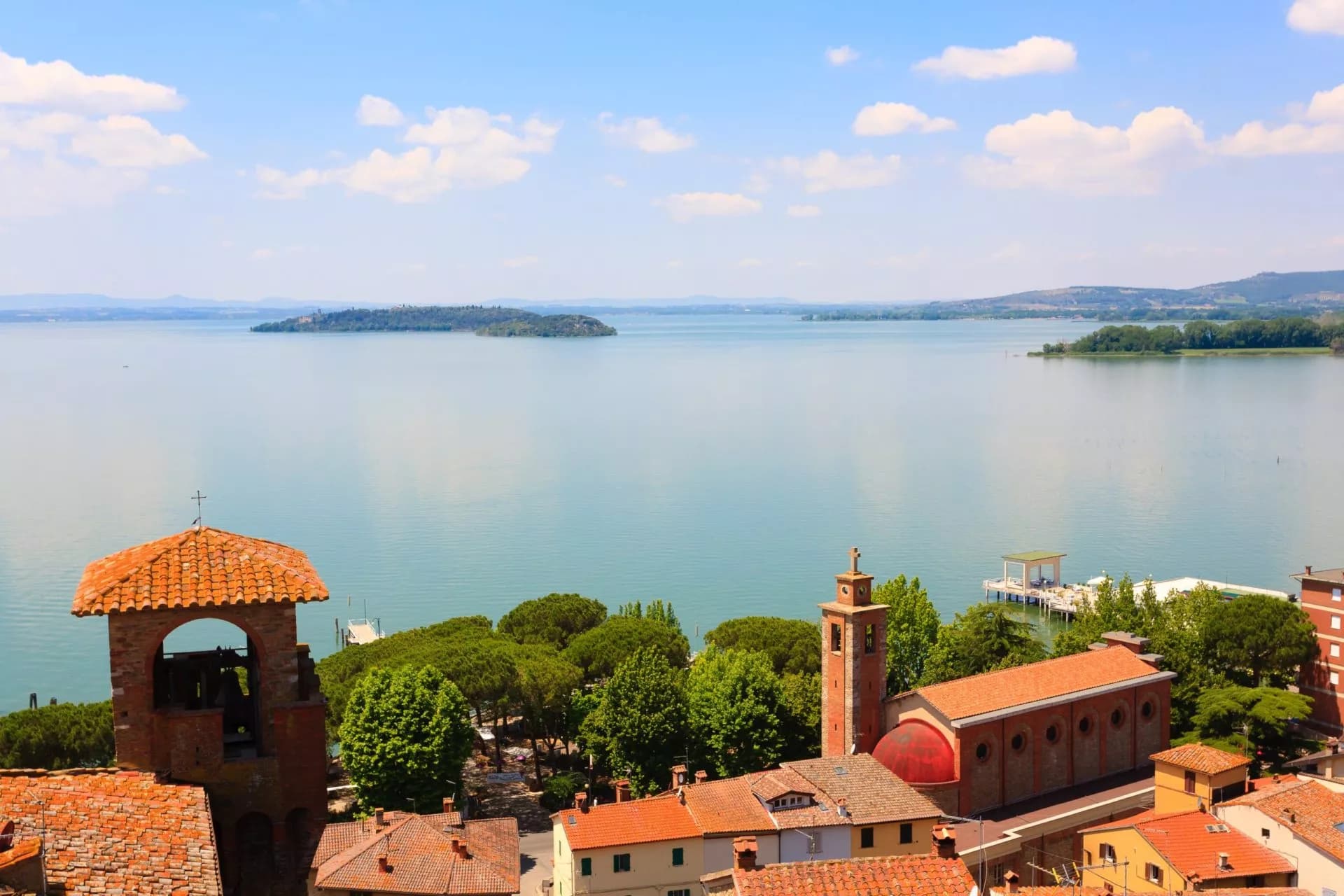 Terracotta roofs and church towers overlooking Lake Trasimeno with islands under a blue sky.