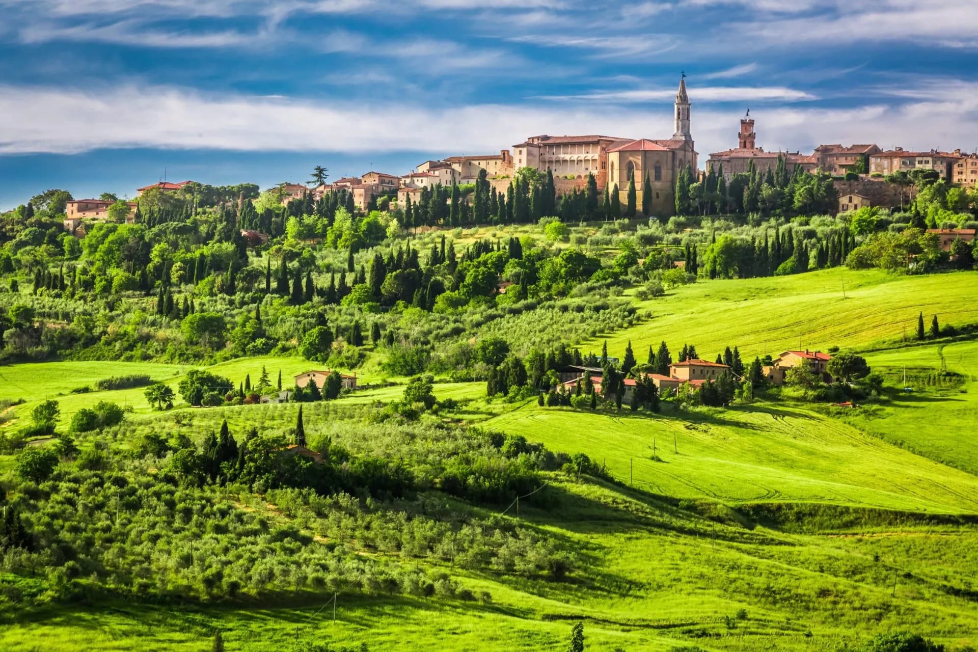 Hilltop town of Pienza with historic buildings above rolling green Tuscan landscape and cypress trees.