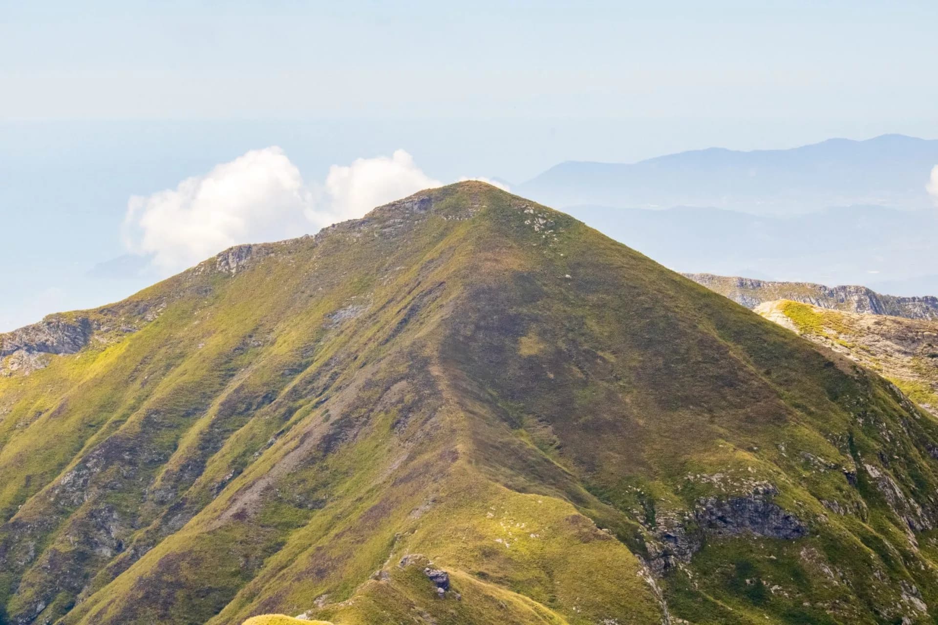 Grassy mountain peak with steep slopes under a pale blue sky, Monte Sumbra visible.