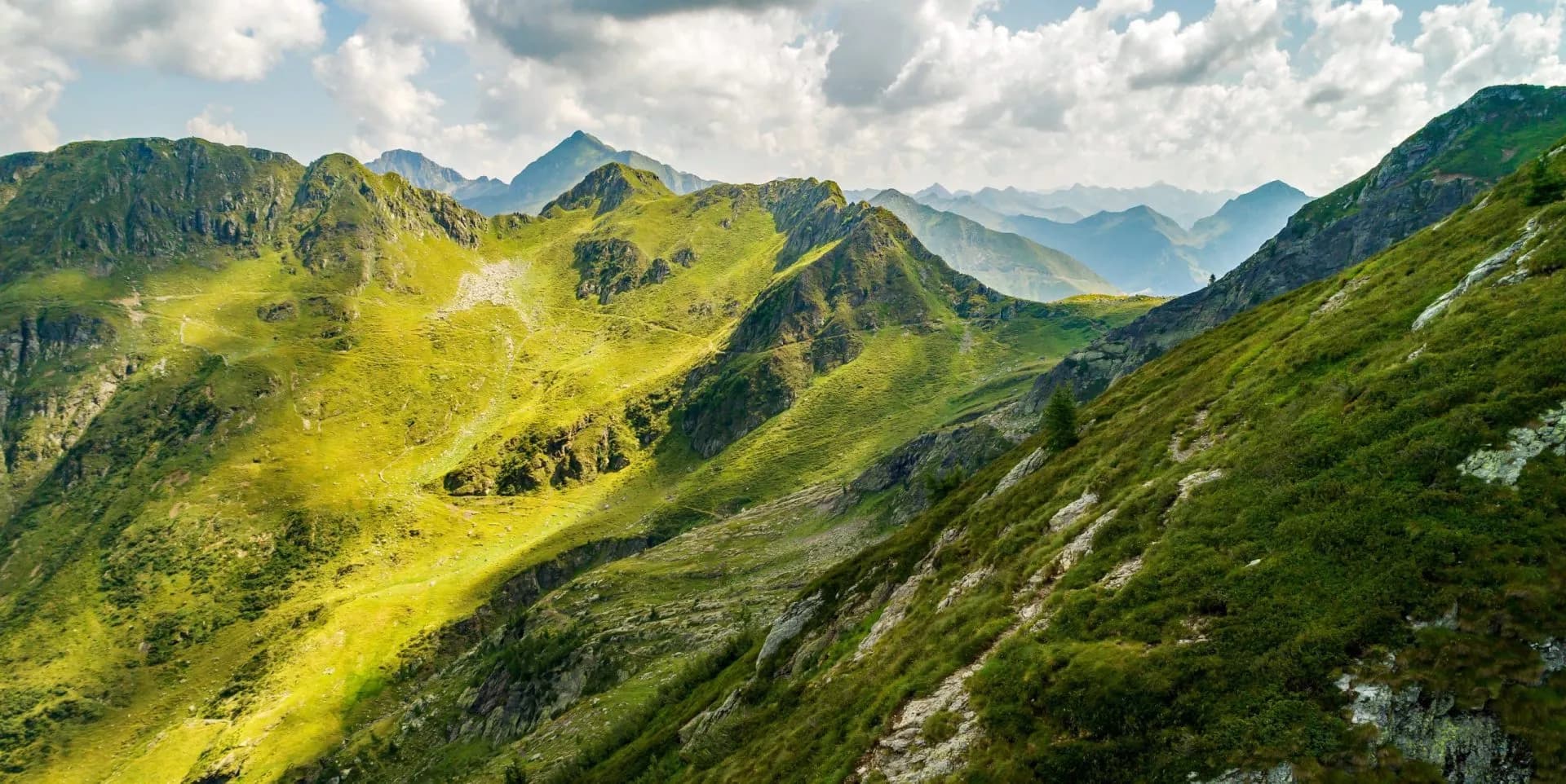 Green, grassy mountain ridges under a cloudy sky, with distant peaks visible
