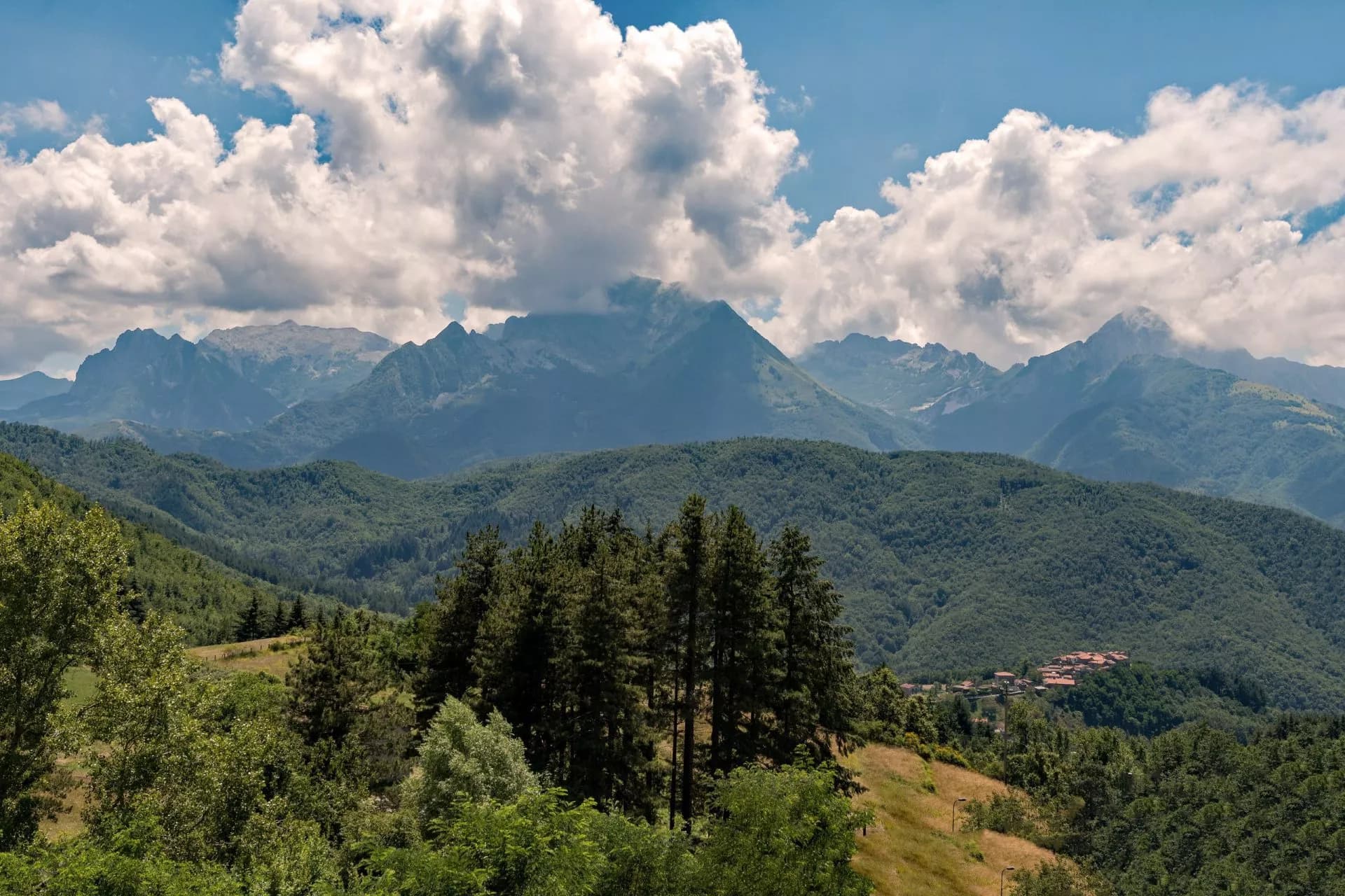 Mountain landscape with lush green hills, pine trees, and dramatic clouds over distant peaks near Casone.