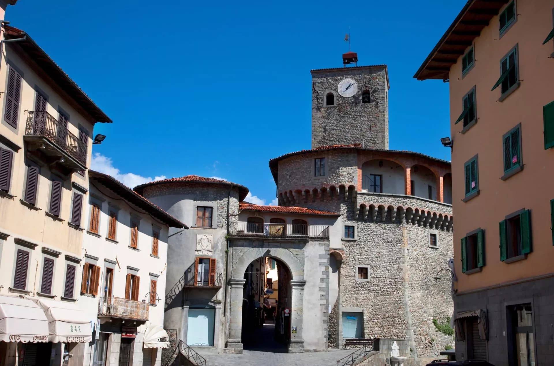 Clock tower and stone archway entrance in Castelnuovo town square under bright blue sky.