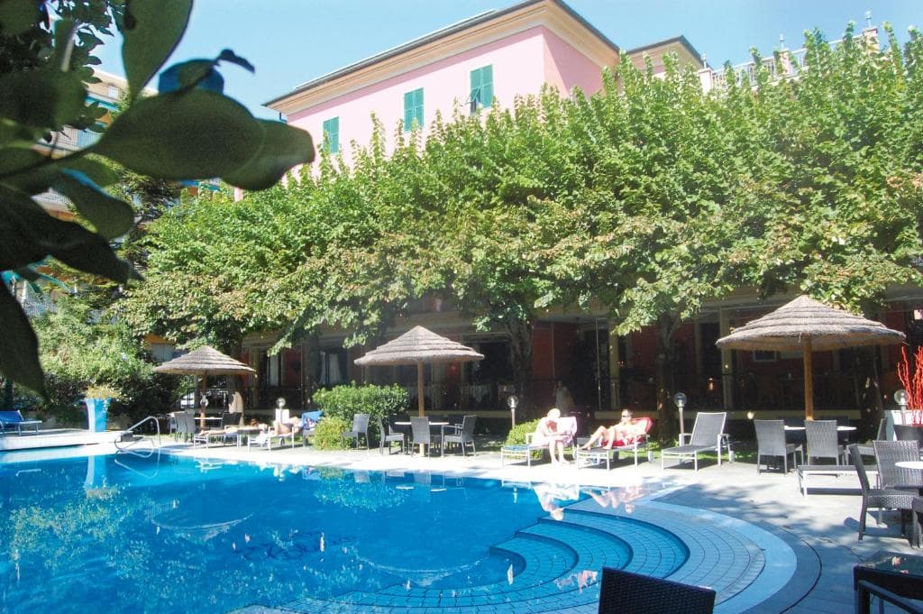 Swimming pool area with sun loungers and thatched umbrellas next to a pink hotel building.